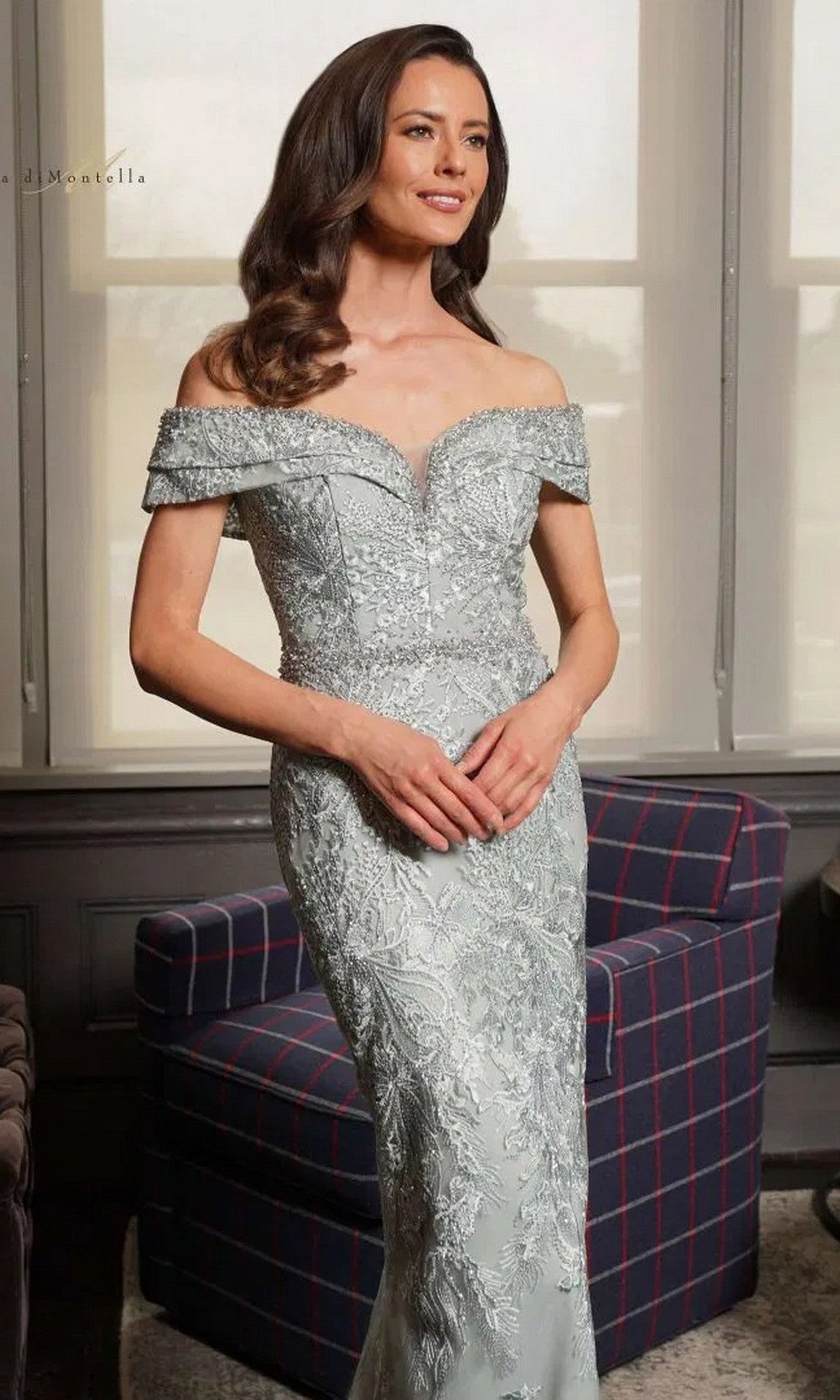 A woman stands indoors before a window and plaid chair, wearing the Rina Di Montella RD2737 long formal dress with an off-the-shoulder neckline, sheer lace details, and a beaded waist.
